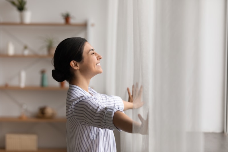 Happy woman open window drapes close eyes in delight.