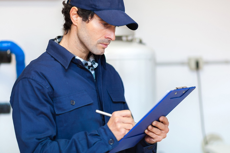 Plumber at work wearing a blue uniform & hat writing on a clipboard, Benefits of Fall Heating Maintenance