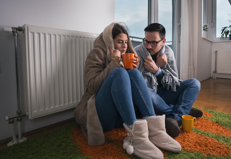 Young couple in jacket and covered with blanket sitting on floor beside radiator and trying to warm up