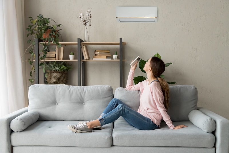 Happy woman rests on couch in modern cozy living room looks up on wall holds cooler system remote controller air conditioner user enjoy fresh air in hot summer day, set comfortable temperature indoors