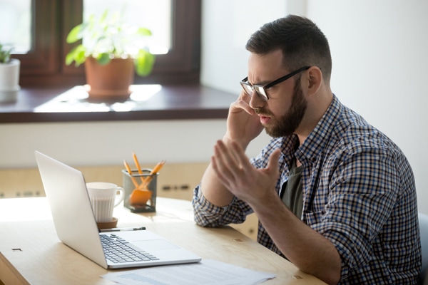 Frustrated male employee discussing his heat pump freezing up over the phone. He is looking at documents in his laptop, holding phone, fling arms in an angry gesture, trying to find solution.