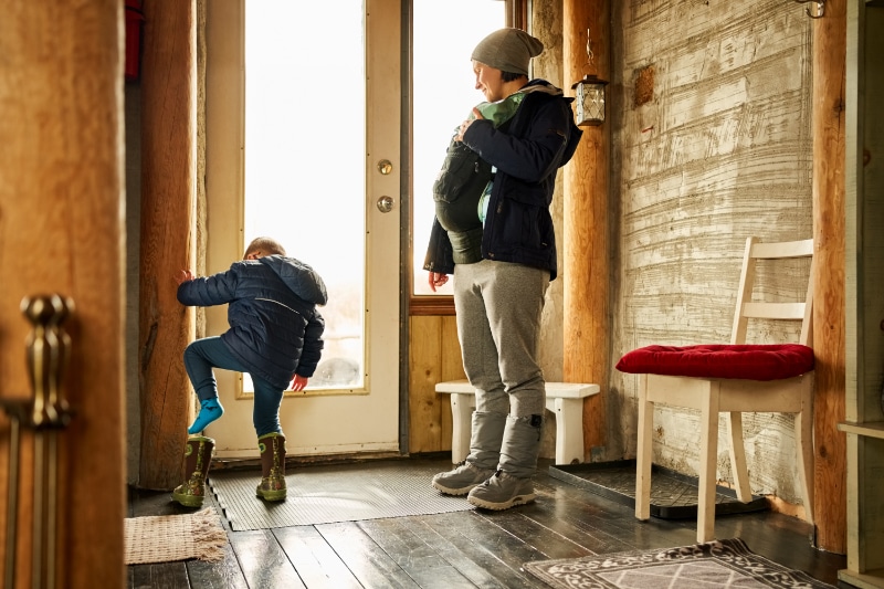 Small boy putting on his boot with his mother standing by carrying a baby boy at front door of eco cabin.