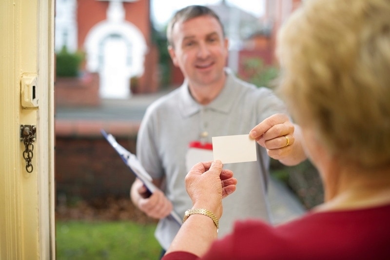 Technician-with-business-card A male technician wearing a gray shirt is handing a business card to a blonde woman in the doorway of her house wearing a red top, Questions to Ask a Plumber